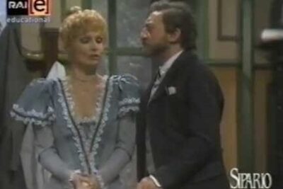 Two actors in period costumes perform a tense, expressive stage scene that visually suggests libero scambio.