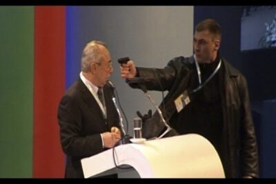 Man aims a handgun at a suited speaker at a podium with microphones confronting lideres politicos
