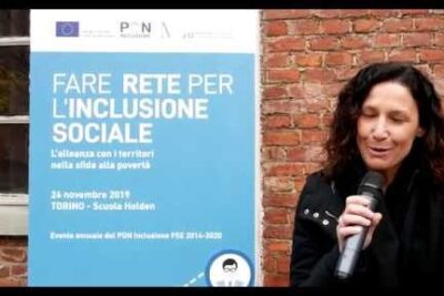 Woman speaking into a microphone beside a blue poster promoting linclusione sociale at a public event.