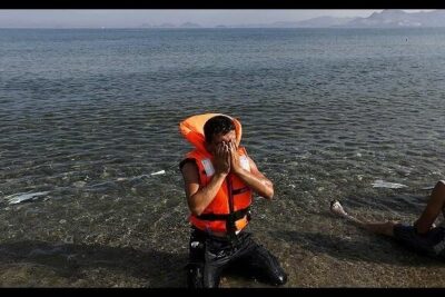 Man kneeling on a rocky shore wearing an orange life jacket and covering his face during llegada inmigrantes 1