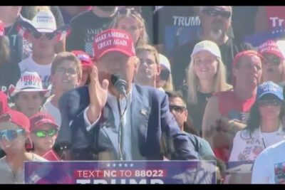 Speaker at an outdoor campaign rally addressing a crowd of supporters wearing red caps, flags and signs — los debates 1