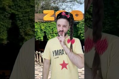 Man in a yellow t-shirt holding a finger to his lips in front of a zoo sign, Playful Visitor at los zoologicos 2 Entrance with Braided Bow.
