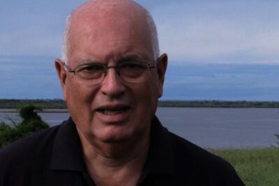 Close-up of a bald man wearing glasses at a lakeside under a cloudy sky, los zoologicos 4.