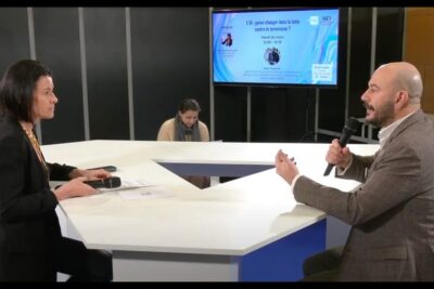 Two people at a studio roundtable speaking into microphones with a display about lutte terrorisme visible behind them.