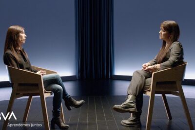 Two women seated facing each other in a minimal studio set for an intimate motherhood and parenting talk, maternidad crianza 1.