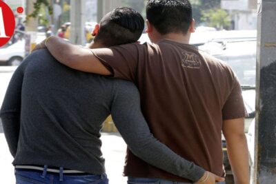 Two men walking closely arm in arm on a sunlit sidewalk, intimate moment representing matrimonio gay 2