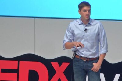 A casually dressed speaker on a TEDx stage holding a remote with a lapel microphone, ideal for media social promotion.