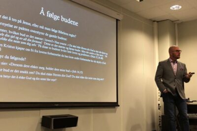 Presenter standing beside a projected Norwegian lecture slide in a seminar room discussing menneskets rolle 1.