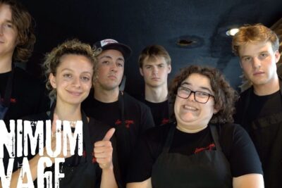 Six young pizzeria employees in black shirts and aprons posing together beneath a 'Minimum Wage' graphic referencing minimum wage 1.
