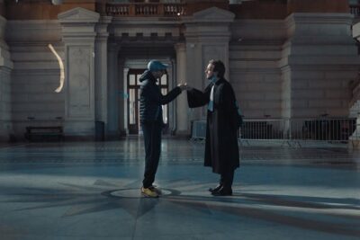 Two people exchange a fist bump in a large marble hall illustrating contrasting mode vie and clothing styles.