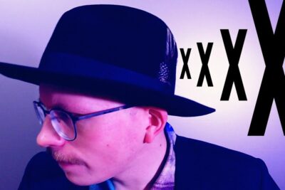 Side-profile portrait of a man wearing a fedora and glasses with large black X graphics on a purple background, modernin taiteen.