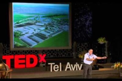 Presenter on a TEDx Tel Aviv stage with a large projected aerial image of agricultural facilities referencing modificerede organismer 1.