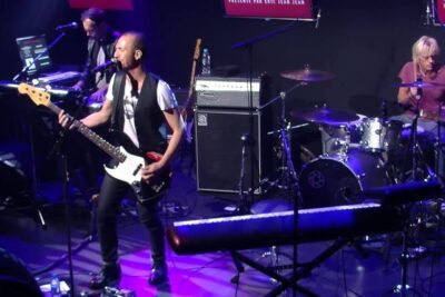 Bassist singing on stage with drummer and keyboardist under colorful lights, reflecting a vibrant monde moderne live performance.
