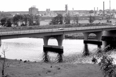 Bridge spanning a river with monsanto on industrial buildings visible in the background.
