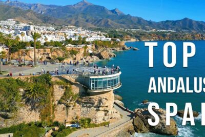 Glass-walled cliffside observation deck overlooking turquoise sea, white coastal town, and mountains, a striking monumenter steder and scenic viewpoint.