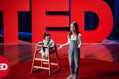 Two children on a red TED stage, one speaking with open hands and another seated in a wooden high chair, illustrating muligheten a 1.