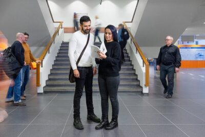 Two people standing in a modern lobby reading a brochure by a central staircase muutos hyvaksi 1