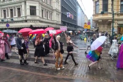 Crowd marching under colorful umbrellas and rainbow flags in a rainy city parade featuring myos niita.