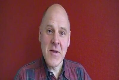 Headshot of a bald man wearing a plaid shirt against a red textured wall for narvarande skolor 1