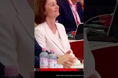 Woman in a white blazer seated at a conference table with bottled water and laptops, illustrating nationalistische haltung.