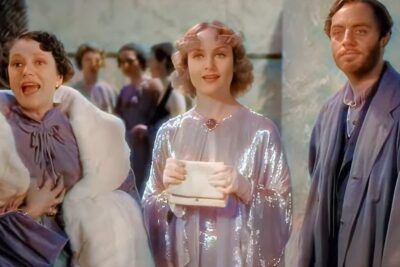 Three performers in vintage costumes onstage with a shimmering-gowned woman holding a clutch between an excited companion and a thoughtful man, nogle mener.