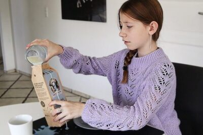 Girl in a lavender knit sweater pouring a pale liquid into a carton at a kitchen table, nogle mennesker