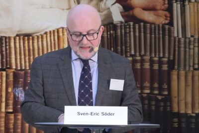 Speaker at a lectern in front of a bookshelf backdrop discussing obligatorisk vaccination