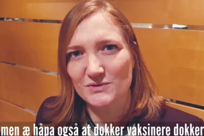 Close-up portrait of a woman with reddish hair against wooden slats with an on-screen subtitle referencing obligatorisk vaksinering 1
