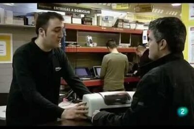 Two men at an electronics service counter with a printer and laptops on shelves, depicting obsolescencia programada 3.