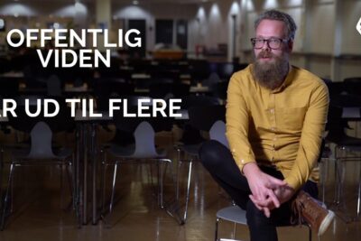 Bearded man in a yellow shirt sits in an empty meeting room illustrating offentlige virksomheder outreach.