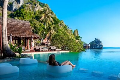 Person reclining on a floating seat in a turquoise infinity pool at a tropical resort with palm trees and ocean islet, okad befolkningsokning.