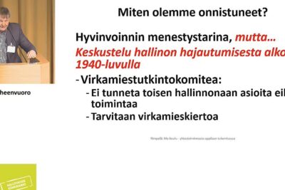 Presenter at a lectern with a Finnish slide about wellbeing and administrative decentralization impacting olevien oppilaiden.
