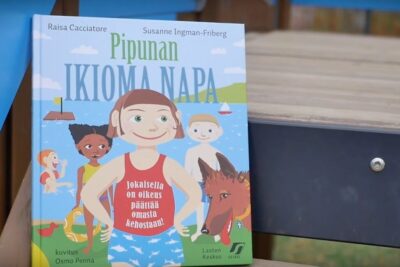 Colorful illustrated children's book cover with smiling kids, a dog and seaside motifs featuring the text omasta kehostaan 1
