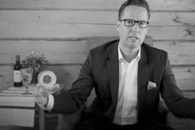 Man in a suit and glasses seated in an armchair gesturing with hands in a rustic wooden room beside a table with a wine bottle and glass, omistaminen voi 1.