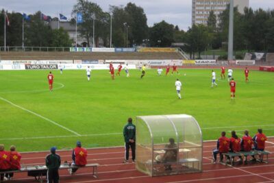 Players in red and white kits contest a soccer match on a green pitch with coaches and substitutes on the sideline on tarkeaa 13.