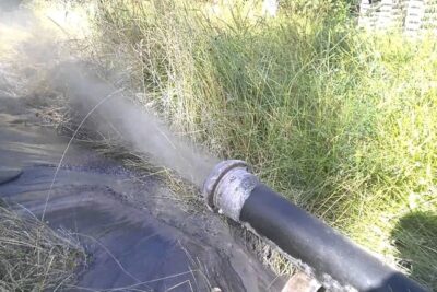 A black metal pipe releasing a forceful muddy water spray into a grassy embankment on tarkeaa 73.