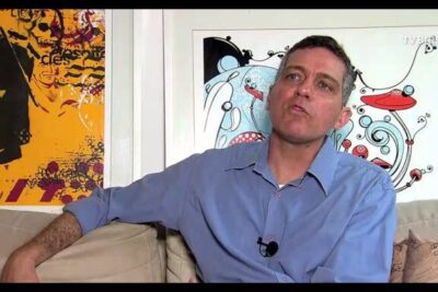 Man in a blue shirt seated on a couch with abstract artwork behind him, implying opinioes divergentes.