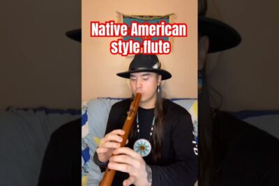 A musician in a hat and beaded necklace playing a wooden Native American style flute showcasing oprindelige folk musical tradition.