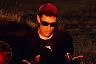 Man wearing sunglasses and dark jacket gesturing in a red-lit field evoking an oui non moment.
