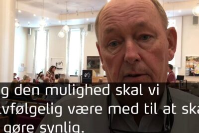Close-up of a man speaking in a community meeting about pa arbejdsmarkedet with audience and Danish subtitles visible