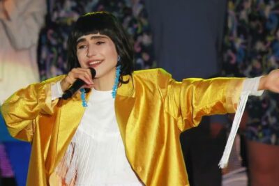 Singer in a gold jacket holding a microphone with outstretched arms and blue earrings pa jorden 8