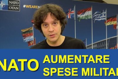 Person standing before NATO logo and multiple national flags with a banner referencing paesi membri and military spending