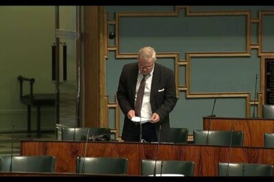 Paljon keskustelua 1: man in a dark suit and tie reading papers at a lectern in a wood-paneled meeting room with empty seats and microphones