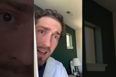 A clinician in a white coat peeks around a doorway in a home interior with a lamp and green accent wall, conveying concern and attentiveness for palliativ omsorg 1