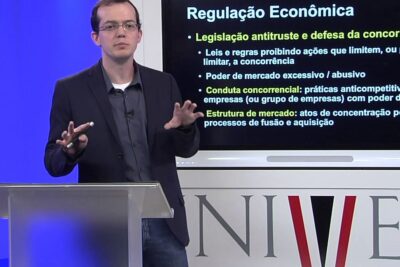 Lecturer at a podium presenting a slide on Regulação Econômica that highlights papel governo and antitrust concepts.