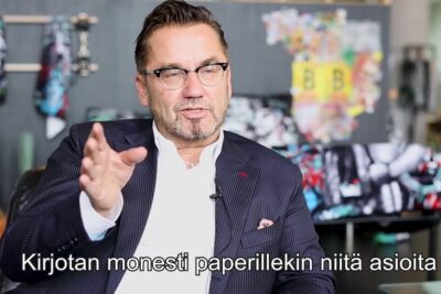 Middle-aged man in a striped suit and glasses gesturing during an interview in a studio with colorful artwork and Finnish subtitles, parantaa ihmisten 1.