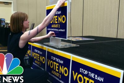 Person arranging 'Stop the Ban — Vote No' signs at a campaign table supporting partisans lavortement.