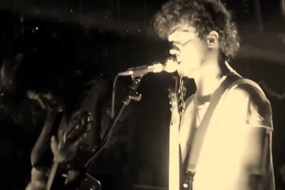 Sepia-toned live performance of a singer and guitarist under dramatic stage lighting, passato migliore