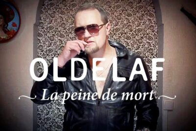 Man in leather jacket and sunglasses posing before patterned wallpaper with prominent text including peine mort 7