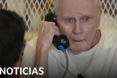 Elderly inmate in a white jumpsuit speaking on a prison phone behind a mesh partition, pena muerte 19